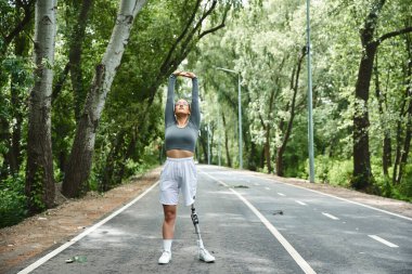 A young woman in sportswear stretches in nature, utilizing her prosthetic leg as she enjoys the outdoors. clipart