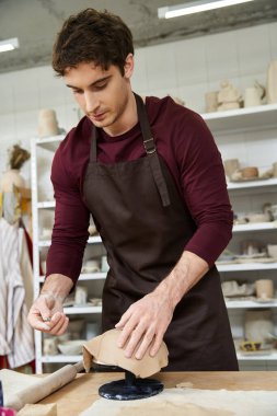 Young man with dark hair making pottery in well lit studio. clipart