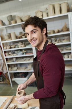 Joyous handsome man in apron making some pottery in studio. clipart