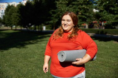 Kendine güvenen bir kadın, canlı yeşil bir parkta yoga minderi tutarken gülümsüyor..