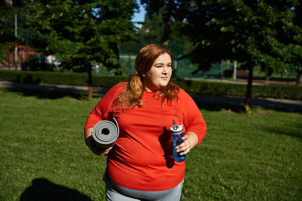 Живучая женщина с большими размерами несет свой коврик и бутылку с водой, готовая тренироваться на свежем воздухе.