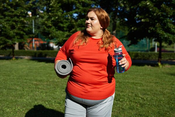 A joyful plus size woman carries a mat and water bottle while exercising outdoors.