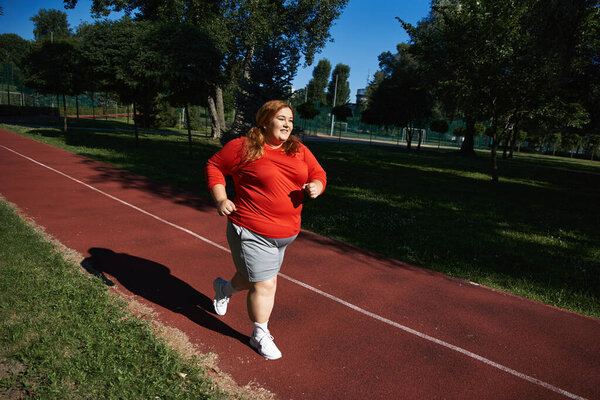 Plus size woman jogs happily along a park path under a clear blue sky.
