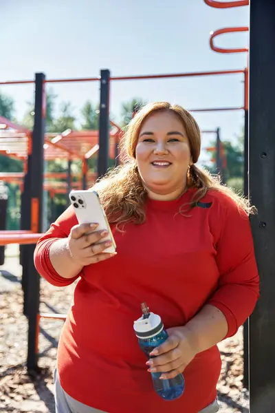 stock image A happy plus size woman exercising outdoors, holding her phone and water bottle.