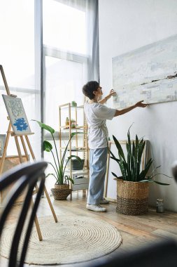 A mature woman curates her art in a sunlit studio filled with plants. clipart