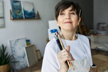 Bir ressam, canlı sanat stüdyosunda elinde fırçalarla gülümsüyor..