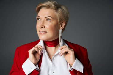 Sofistike giyinmiş yetişkin bir kadın yakasını düşünceli bir bakışla düzeltir, zarafet saçar..