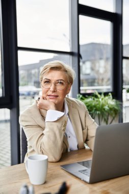 Gözlüklü zarif bir kadın şık bir ofis masasında otururken işini düşünüyor..