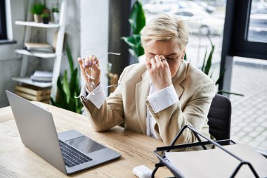 Şık giyinen kibar bir kadın, öğle vakti modern ofis masasında düşünceli bir şekilde duruyor..