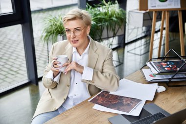 A confident woman in chic attire sips coffee at her stylish desk, surrounded by plants. clipart