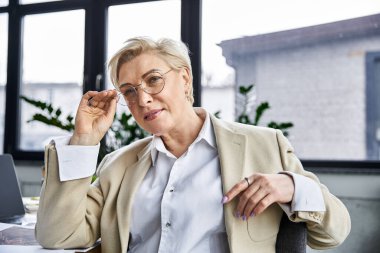 A stylish woman adjusts her glasses while pondering ideas in a modern office. clipart