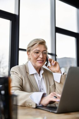 Şık bir kadın zarif bir mekanda dizüstü bilgisayarı üzerinde çalışırken telefonuyla sohbet ediyor..