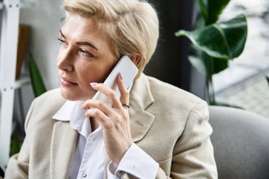 Şık giyinmiş şık bir yetişkin kadın ofiste otururken telefonla iletişim kuruyor..