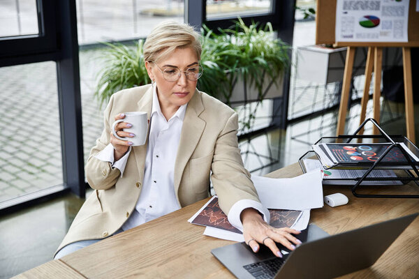 Элегантная взрослая женщина пьет кофе, сосредоточившись на ноутбуке..