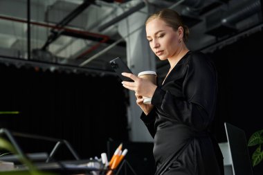 Genç, güzel, iri yarı bir kadın iş yerinde kahvenin tadını çıkarırken akıllı telefonuna odaklanıyor..