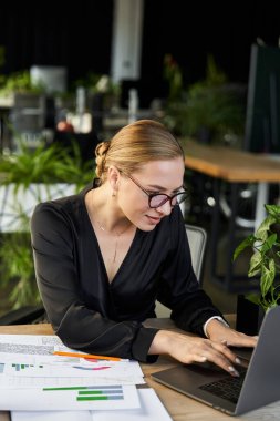 Genç ve güzel bir kadın laptopuna odaklanıyor. Etrafı canlı ofis bitkileriyle çevrili..