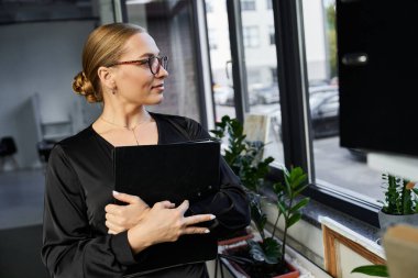 A confident young woman reflects thoughtfully while holding a folder in her office workspace. clipart