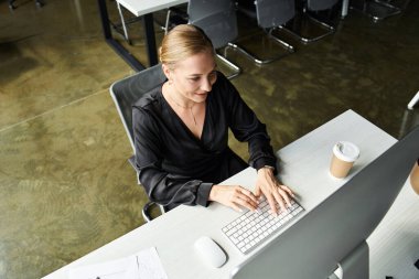 A beautiful young woman types attentively at her desk, savoring her coffee during work hours. clipart