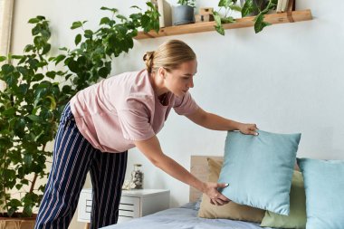 A young plus size woman arranges colorful pillows on her tidy bed among lush greenery. clipart