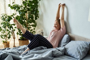 A cheerful young woman stretches happily in her inviting bedroom, radiating positivity. clipart