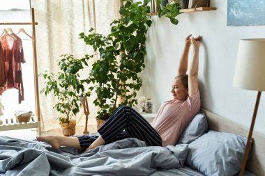 In a bright and inviting room, a young beautiful woman stretches joyfully in bed. clipart