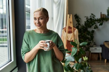 A young beautiful plus size woman stands by the window, savoring her warm drink in comfort. clipart