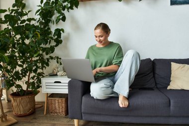 A young woman relaxes at home, engaged with her laptop in a stylish living room. clipart