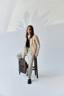 Şık bir genç adam bir taburede duruyor, modaya uygun giysiler ve parlak bir çanta sergiliyor..