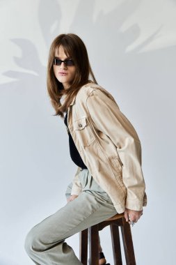 Şık bir genç, minimalist bir ortamda şık bir kıyafet sergileyerek bir taburede poz veriyor..