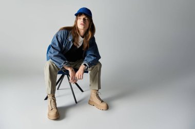 Genç bir adam minimalist bir ortamda sandalyede otururken modaya uygun kıyafetlerini sergiliyor..