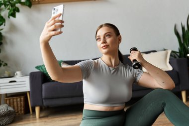 Kırpılmış bluz ve tayt giymiş sportif bir kadın selfie çekerken ev egzersizi yapıyor..
