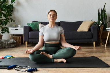 Sportif genç bir kadın evdeki paspasın üzerinde meditasyon yapıyor. Etrafı spor aletleri ve rahat bir çevreyle çevrili..