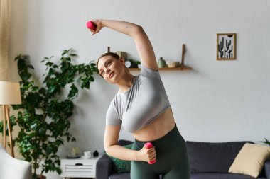 Tozlu ve ekinlerle evde spor yapan genç bir kadın, antremanlarını geliştirmek için dambıl kullanıyor..