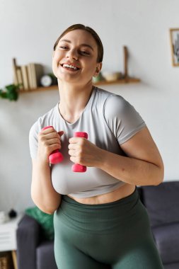 Genç, sportif bir kadın evde egzersiz yaparken gülümsüyor, şık spor kıyafetleriyle pembe ağırlıklarını kaldırıyor..