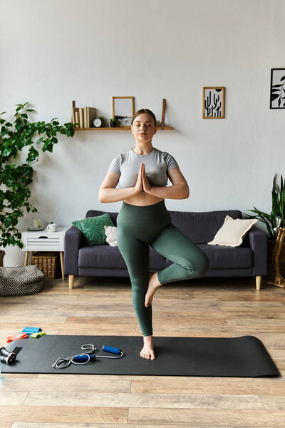 Молодая женщина в активной одежде балансирует на одной ноге во время занятий йогой, создавая спокойную атмосферу дома.