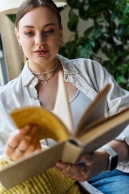 Günlük kıyafetli genç bir kadın evinde rahatça otururken kitap okuyor..