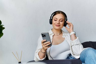 Gündelik kıyafetli genç bir kadın akıllı telefonuyla aktif olarak uğraşırken kulaklıkla müzik dinliyor..