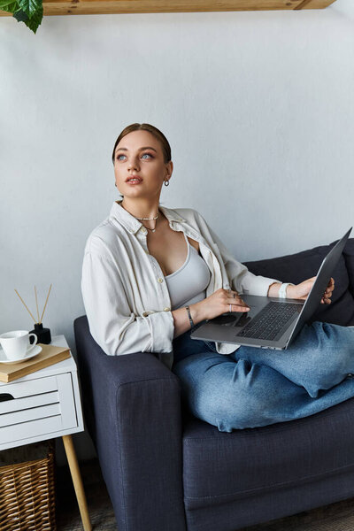 Молодая женщина, небрежно одетая, удобно сидит на диване и пристально работает над ноутбуком..