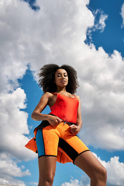 Стильная молодая женщина гордо стоит в ярком одеянии, излучая уверенность под голубым небом.