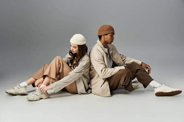 A fashionable couple sits back to back, showcasing autumn attire and trendy footwear.