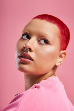 Cesur genç bir kadın benzersiz tarzını sergiliyor. Kısa pembe saçları ve parlak bir arka planda burun piercingiyle..