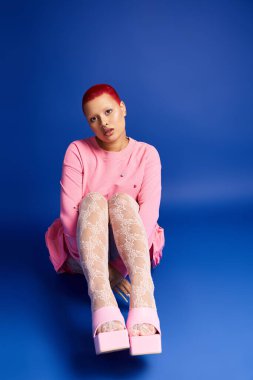 Pembe saçları ve burun piercingleri olan genç bir kadın stüdyoda kendine güvenerek oturuyor ve kendine has tarzını benimsiyor..