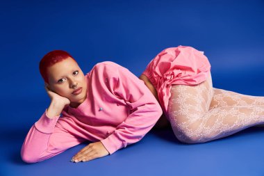 Pembe saçlı cesur genç bir kadın, kendine güveni tam, canlı, mavi bir arka plana karşı eşsiz tarzını sergiliyor..