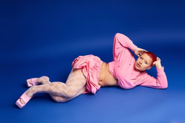Pembe saçlı ve burnuna piercing takmış kendine güvenen genç bir kadın mavi bir arka plana şık bir şekilde poz verir..