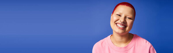 Веселая молодая женщина с короткими розовыми волосами и пирсингом носа выражает свою смелую личность в яркой студии.