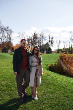 Sevgi dolu bir çift, canlı yapraklar ve güneş ışığıyla çevrili sonbahar kıyafetleriyle rahat bir yürüyüş yapar..
