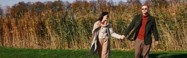 A loving couple walks hand in hand, surrounded by vibrant autumn foliage under clear skies. clipart