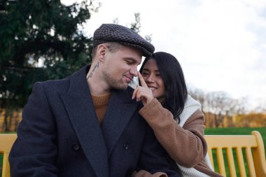 Bir çift, sonbahar kıyafetleri içinde, parkta bir bankta oturmuş, gülümseyerek hassas bir anın tadını çıkarıyor..