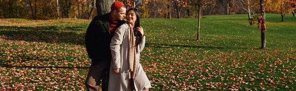 A warm and appealing couple embraces each other under autumn trees, surrounded by fallen leaves.