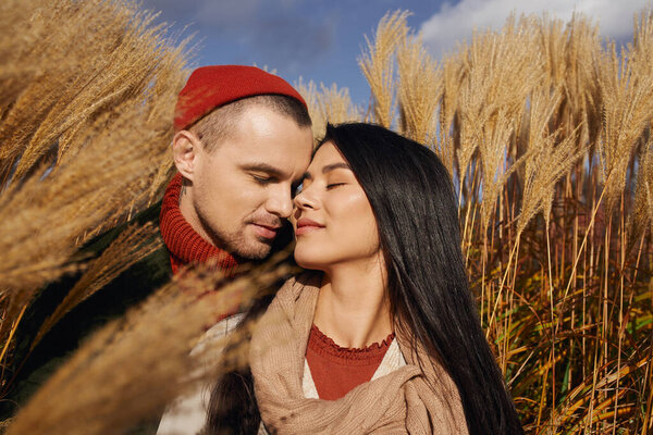 A couple dressed warmly embraces, smiling gently against a backdrop of autumn foliage and grasses.